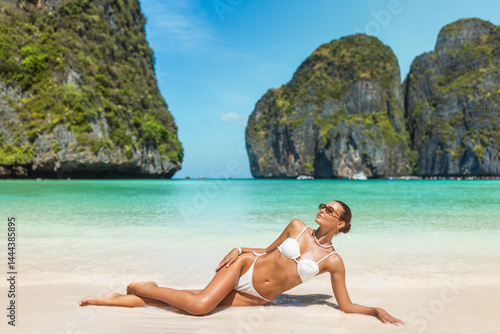 Stunning portrait of an attractive woman relaxing and sunbathing in a white bikini on the pristine sands of Maya Bay, Ko Phi Phi. Surrounded by breathtaking turquoise waters and dramatic limestone