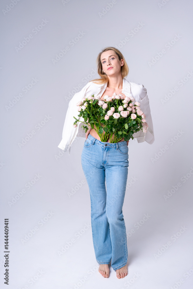Young woman holding a big bouquet of roses standing barefoot in blue jeans and a white shirt in a bright studio. Suitable for joy, celebration and style concepts