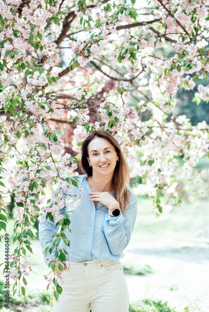 Smiling woman standing among blooming pink cherry blossom sakura trees wearing a light blue shirt and enjoying a bright spring day outdoors Smiling woman standing among blooming pink cherry blossom sakura trees wearing a light blue shirt and enjoying a bright spring day outdoors