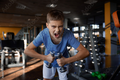 Teenage boy grimacing screaming to camera while training with dumbbells