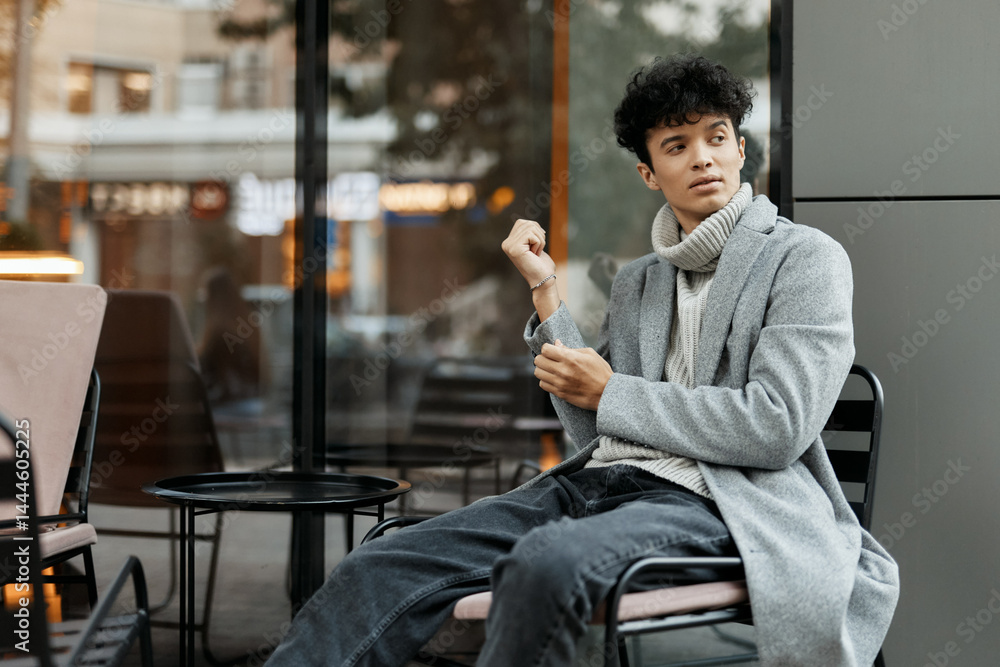 Fashionable young man sitting in chair by window with hands in pockets and cigarette in mouth