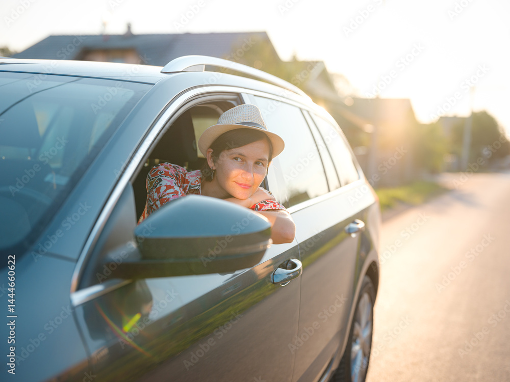 Naklejka premium Traveler woman in red ethnic dress and hat drives car through scenic countryside of Croatia in summer. Surrounded by lush fields and forests, she enjoys peaceful and adventurous road trip