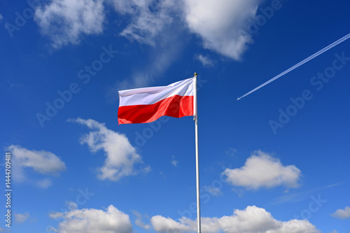 The polish flag flutters in the wind an aeroplane flies past at high altitude with a contrail