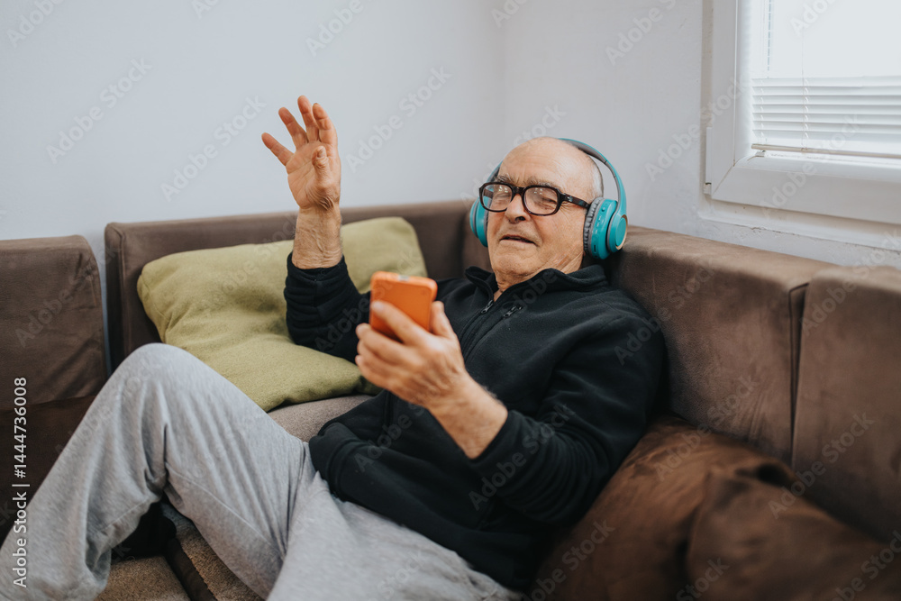 A senior man enjoying music through headphones while using a smart phone, reclining on a comfortable couch in a living room setting with soft pillows and sunlight filtering through a window.