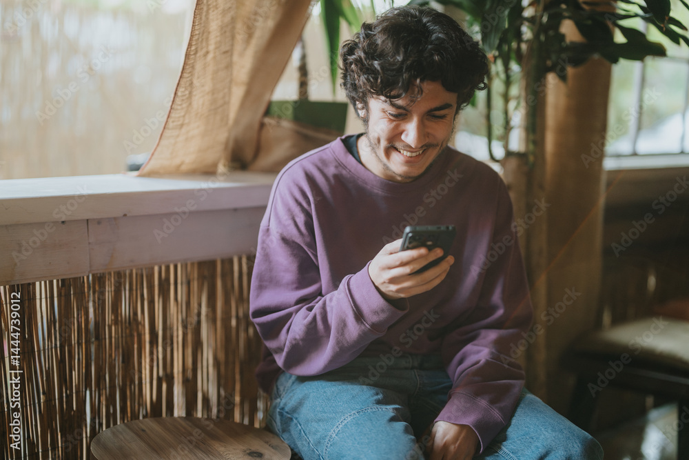A young man wearing a purple sweater sits in a cozy environment, smiling as he looks at his smart phone. The natural lighting and comfortable atmosphere add warmth to the scene.