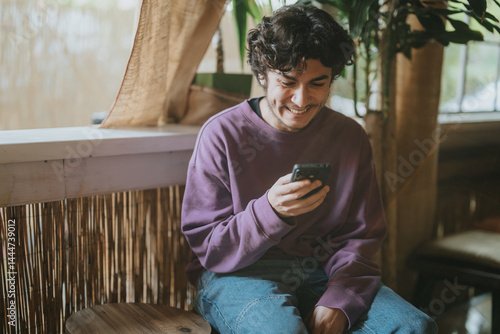Obraz na płótnie A young man wearing a purple sweater sits in a cozy environment, smiling as he looks at his smart phone. The natural lighting and comfortable atmosphere add warmth to the scene. w salonie