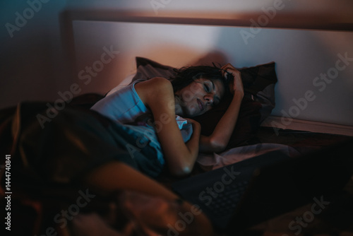 A woman peacefully sleeps in a dimly lit bedroom beside her open laptop, conveying a sense of relaxation and modern digital lifestyle.