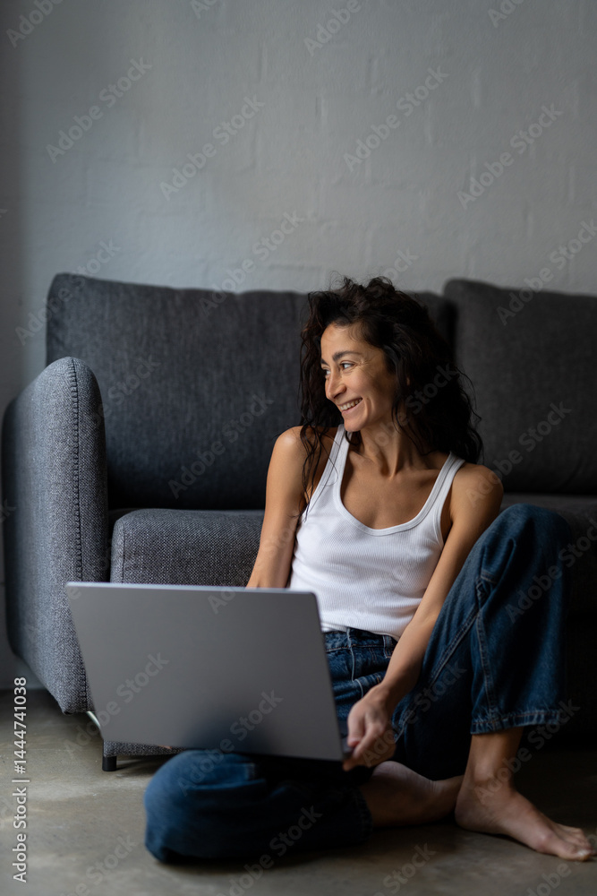 Naklejka premium Happy young freelance woman working from home with laptop, sitting on the floor in casual clothes, smiling and enjoying remote work in cozy relaxed atmosphere.