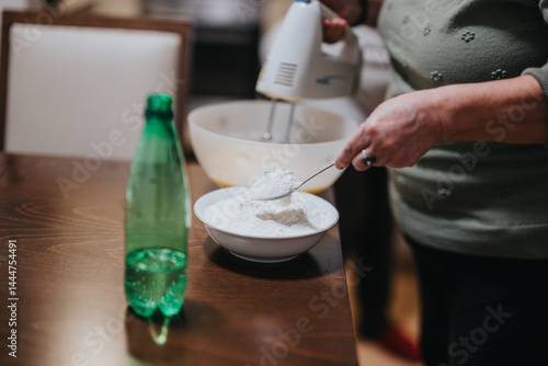 A senior woman is seen preparing a bowl of flour in a kitchen, using a hand mixer and a spoon to combine ingredients while baking homemade food.