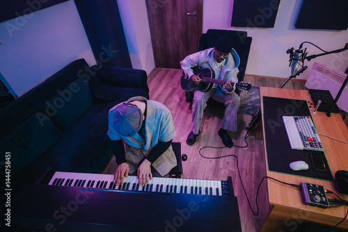 Obraz na płótnie Two musicians, one playing a keyboard and the other a guitar, work together in a contemporary recording studio. The inviting space is filled with musical instruments and professional equipment. w salonie