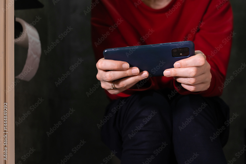 Woman wearing a red sweater and dark pants is holding a smartphone horizontally with both hands, playing a mobile game while sitting on a toilet in a bathroom