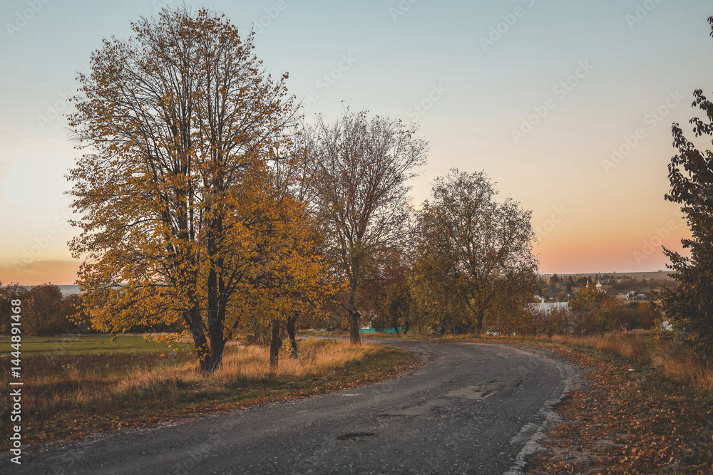 Winding country road through autumn landscape with trees in golden foliage, under a pastel sunset sky in rural countryside Winding country road through autumn landscape with trees in golden foliage, under a pastel sunset sky in rural countryside