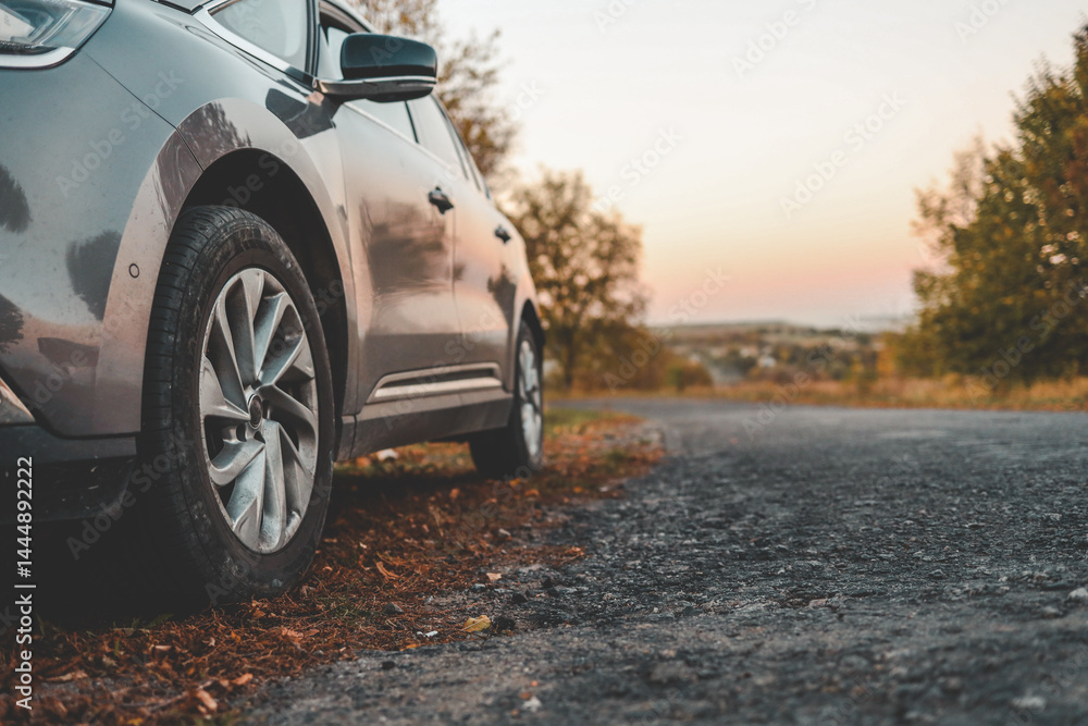 Naklejka premium Close-up of modern car parked on roadside with autumn trees in background. Curved country road leads into soft sunset horizon, evoking peaceful travel mood