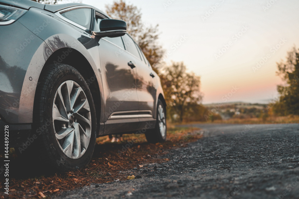 Naklejka premium Close-up of modern car parked on roadside with autumn trees in background. Curved country road leads into soft sunset horizon, evoking peaceful travel mood