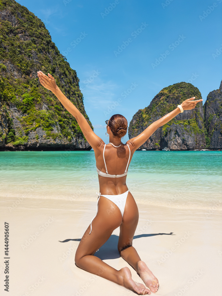 Rear view of tanned woman in white bikini with arms raised joyfully on Maya Bay beach, Thailand - ideal for beauty, lifestyle, tropical travel, vacation freedom and summer beach fashion content.
