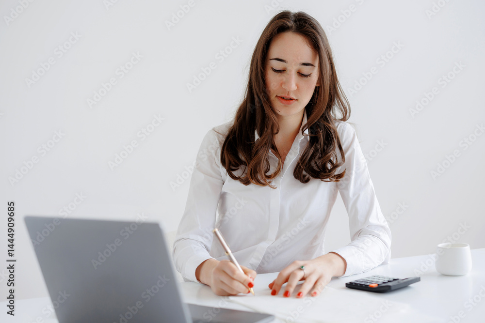 Young woman working diligently at a desk while taking notes and using a laptop in a bright, minimalist office setting