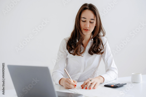 Obraz na płótnie Young woman working diligently at a desk while taking notes and using a laptop in a bright, minimalist office setting w salonie