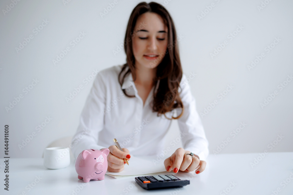 Woman calculating expenses with a calculator and piggy bank on a desk in a bright office setting