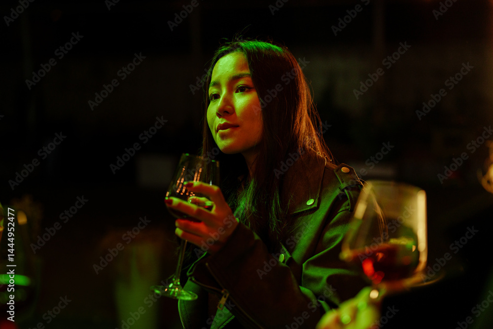 Woman enjoying a drink during a lively gathering in a dimly lit setting with vibrant green lights at night