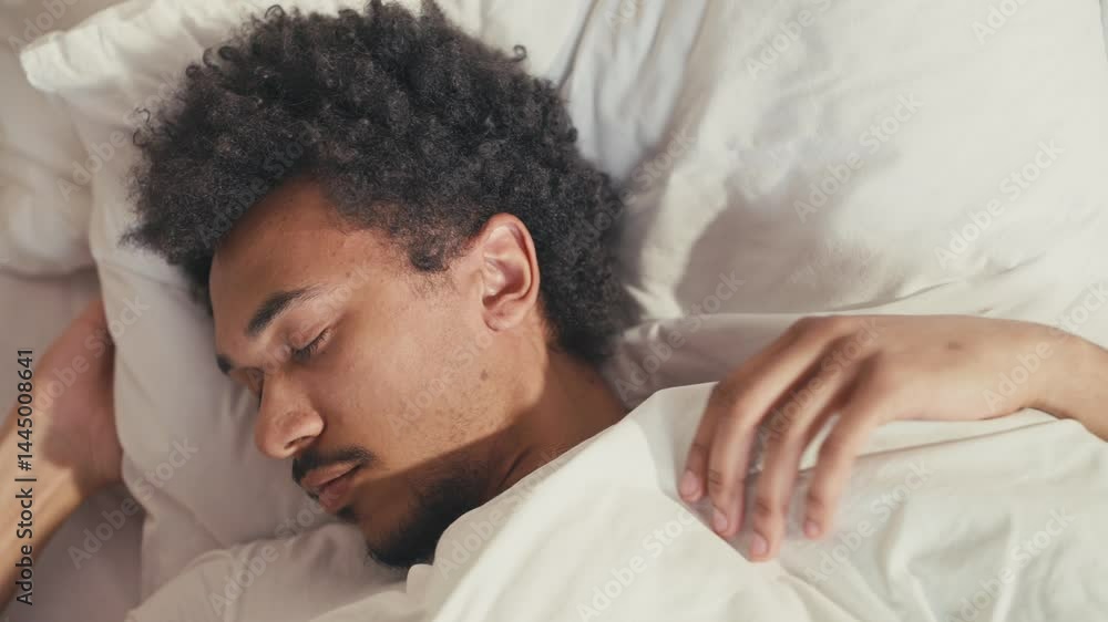 African American male turning over in bed while sleeping. Resting head on white pillow. Wearing white T-shirt, lying under white blanket. Peaceful expression during deep sleep moment. African American male turning over in bed while sleeping. Resting head on white pillow. Wearing white T-shirt, lying under white blanket. Peaceful expression during deep sleep moment.