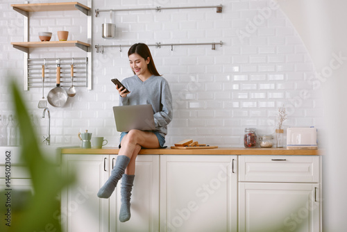 Portrait of beautiful Asian woman using mobile phone while sitting on kitchen counter bar at home. Happy Blogger and freelance work online. Asian young woman with digital technology concept.