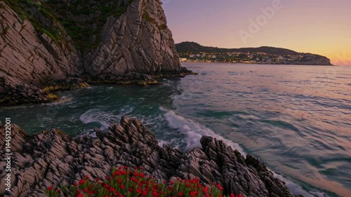 Rugged rocky shoreline at sunset with distant town lights and vibrant flowers