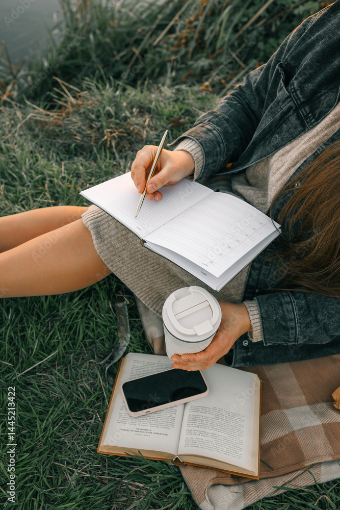 Obraz premium Woman sitting on blanket in grass, writing in a journal with coffee cup in hand. Cozy autumn mood with boots, book, smartphone, and fallen leaves around her