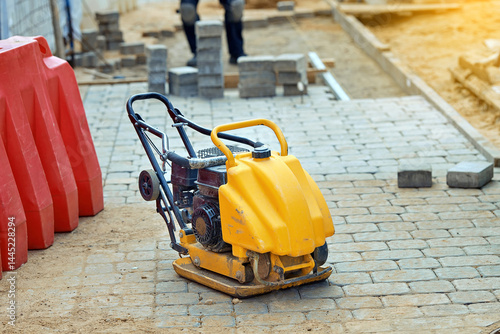 Obraz na płótnie Yellow compactor machine on paved stone walkway at construction site with red barriers and paving slabs. closeup of plate compactor standing on newly laid pavement stones. Vibratory plate compactor w salonie