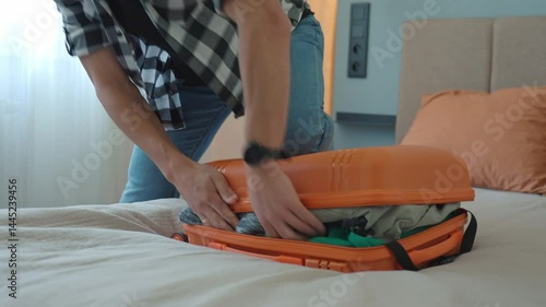 Man trying to close over packed orange suitcase before trip. Male traveler has trouble while packing a lot of clothes, preparing for travel