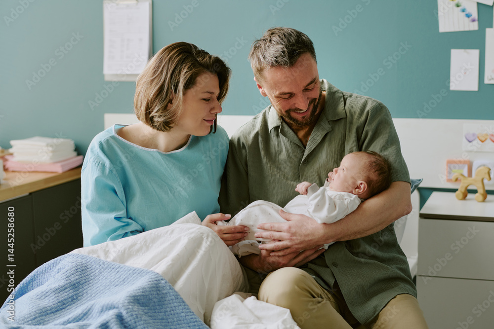 Obraz premium Caucasian parents smiling while holding their newborn baby in a hospital room. The mother sits on the bed covered with a light blanket