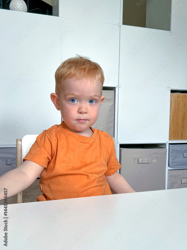 A red-haired boy in an orange T-shirt is sitting at a table in the room. Early development of a child at home