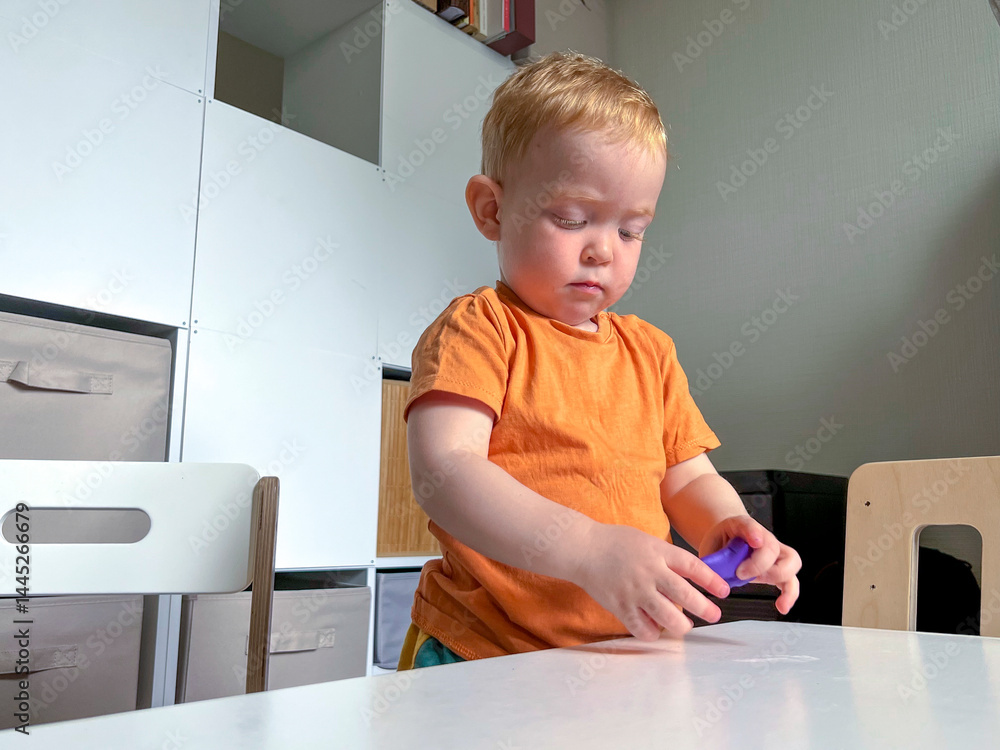 A red-haired boy in an orange T-shirt stands in front of a table and sculpts from plasticine. Early development of a child at home
