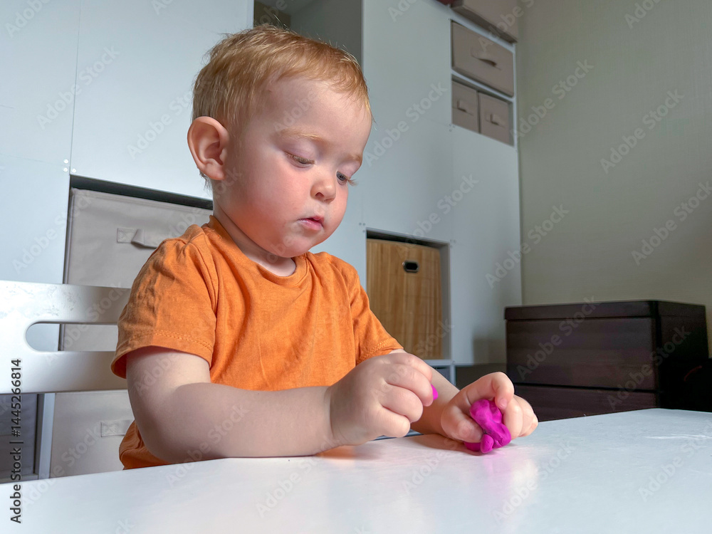 A red-haired boy in an orange T-shirt sculpts purple plasticine at a table. Early development of a child at home
