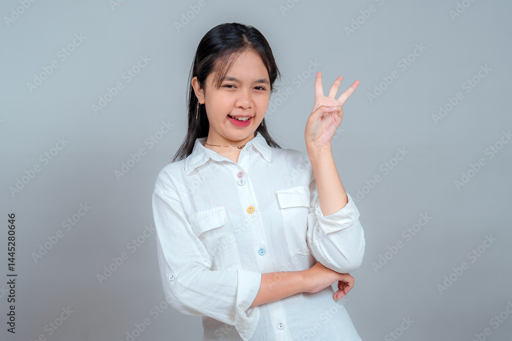 Playful young woman in white shirt making a hand gesture with three fingers up while smiling and winking. Confident expression and fun attitude against a plain grey background. Playful young woman in white shirt making a hand gesture with three fingers up while smiling and winking. Confident expression and fun attitude against a plain grey background.