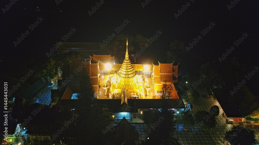Aerial view capturing a brightly illuminated golden pagoda at night, surrounded by intricate temple buildings and lush trees, creating tranquil and spiritual atmosphere. Thailand, Chiang Mai Province
