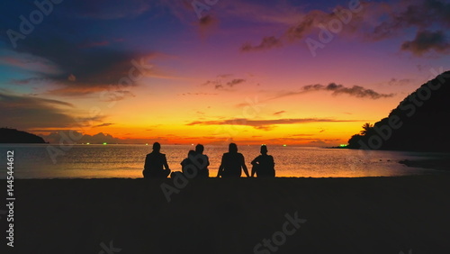 Obraz na płótnie Five tourists sitting on the sandy shores of Koh Ma Beach, Koh Phangan Island, Thailand, are admiring a stunningly colorful sunset painting the sky over the tranquil sea w salonie