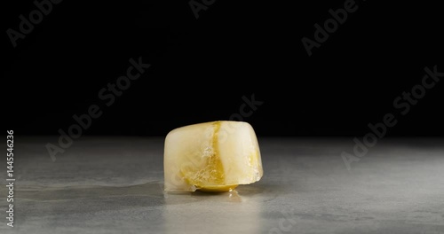 Lemon in an ice cube, close-up. Time lapse of the ice melts and a half of lemon appears. Frozen fruit. Black background.