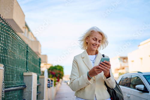 Older woman smiles while using smartphone on sunny street in residential area