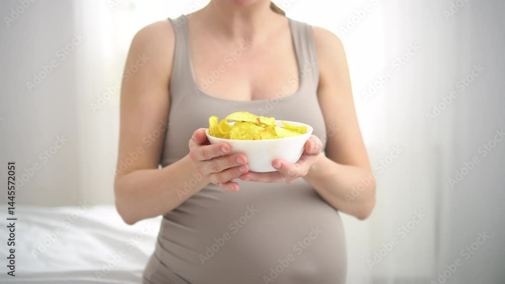 Close-up of future mother snacking on potato chips while stroking her baby bump. Honest portrayal of food indulgences during pregnancy. Close-up of future mother snacking on potato chips while stroking her baby bump. Honest portrayal of food indulgences during pregnancy.