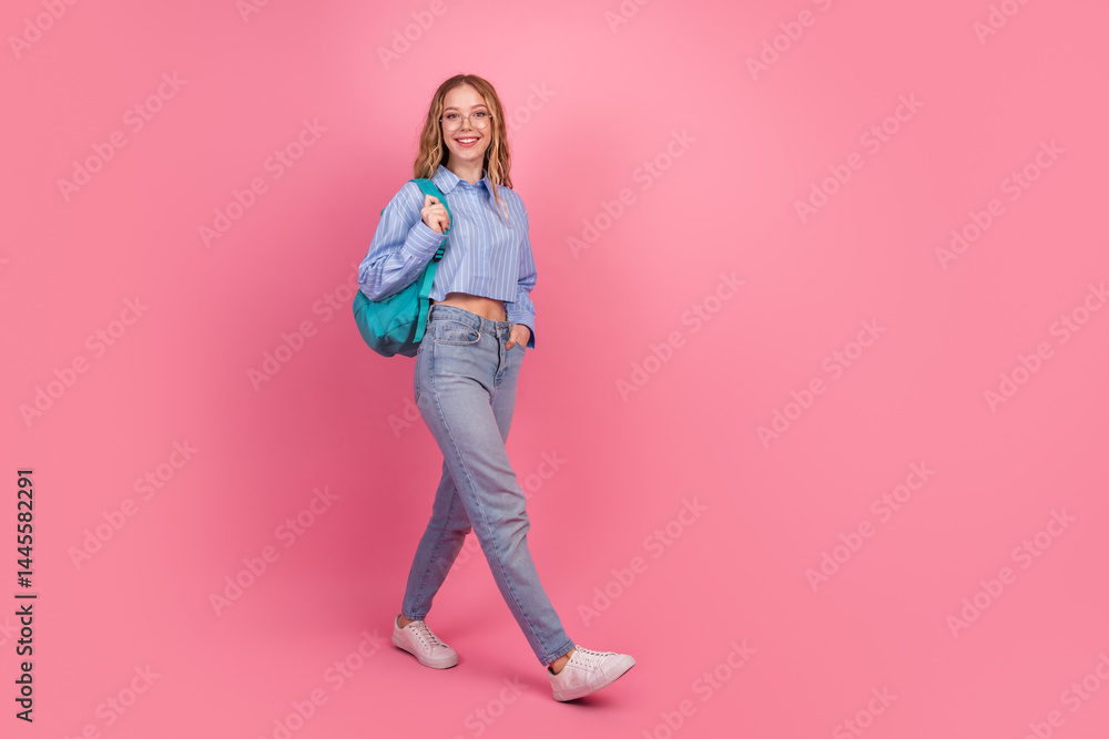Smiling young woman in stylish attire walking confidently with her backpack against a vibrant pink background