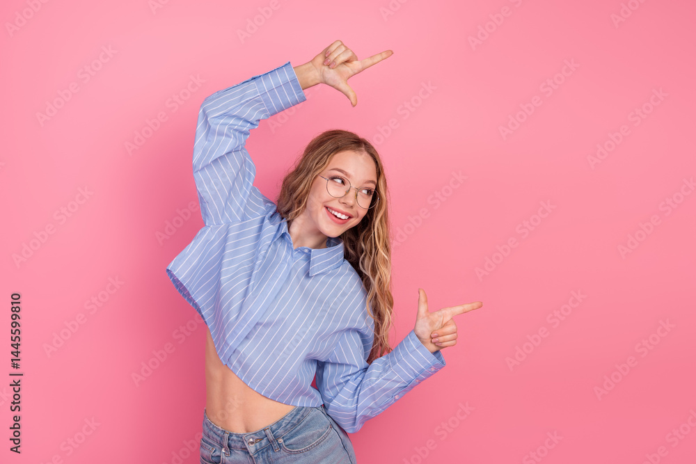 Smiling young woman in a striped blue shirt and casual jeans posing with hand gestures against a pink background.