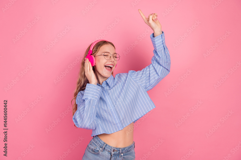 Young joyful woman in stylish attire enjoying music with headphones against a pink background expressing happiness and leisure