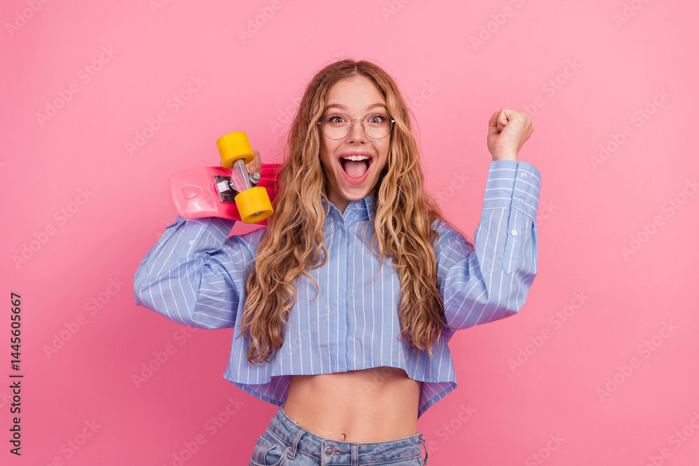 Excited young woman in striped blue shirt holding a colorful skateboard, enjoying leisure time on a pink background
