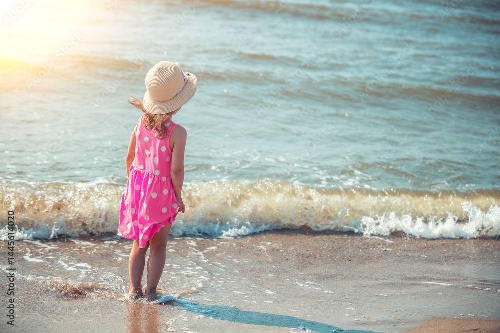 Happy little girl walking on the beach in summer. Child in polka dot dress and straw hat