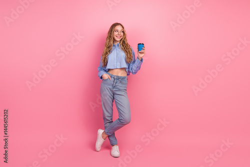 Casually styled young woman with long wavy blond hair in blue outfit holding a cup, smiling in front of pink background
