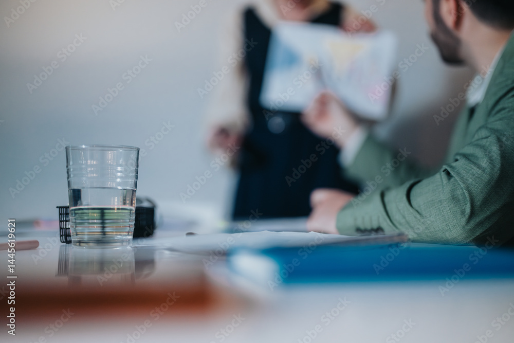 Fototapeta premium A transparent glass of water placed on a desk in an office setting, with a blurred business meeting in the background. Suggests professionalism, focus, and a collaborative work environment.