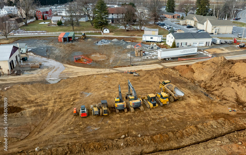 Heavy machinery including excavators and dump trucks is actively working at a construction site in a rural area. Workers are overseeing the excavation process in daylight.