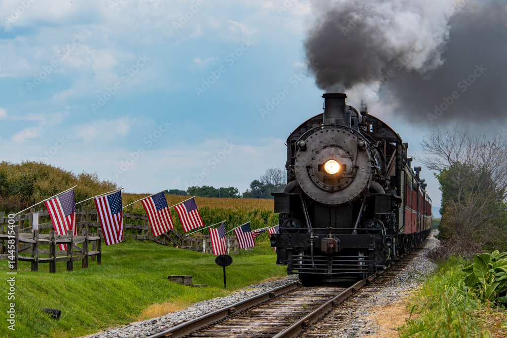Obraz premium A classic steam train makes its way through a picturesque rural landscape, flanked by American flags. The scene captures the charm of vintage rail travel on a clear day.
