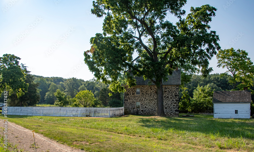 A rustic stone house stands amidst a verdant landscape, featuring a large tree nearby. A white fence frames the property, with a dirt path leading towards the house under clear blue skies. A rustic stone house stands amidst a verdant landscape, featuring a large tree nearby. A white fence frames the property, with a dirt path leading towards the house under clear blue skies.