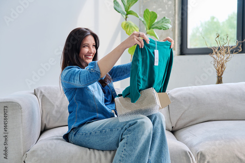 Obraz na płótnie Young woman unpacking cardboard box parcel with online order, looking at new clothes w salonie